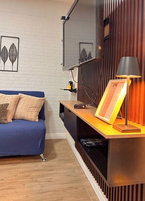 Ett modernt vardagsrum med en blå soffa, ett träbord som fungerar som avlastningsyta och har en lampa på sig, samt en vit tegelvägg.