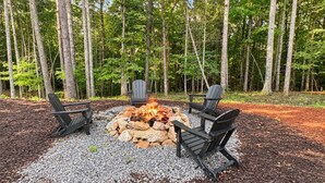 An outdoor seating area with a fire pit surrounded by rocks and gravel, set against a backdrop of trees. - Spur and Arrow - Parade of Homes Cabin | Screened Porch and Hot Tub | Firepit (Morganton)