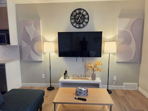 A living room with a flat-screen TV, two wall-mounted lamps, a clock, and a decorative vase with flowers. - NEW 2-bedroom Basement Suite near Airport (Northeast)