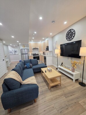 A modern living room with a sectional sofa, a wooden coffee table, and a flat-screen TV mounted on the wall. - NEW 2-bedroom Basement Suite near Airport (Northeast)