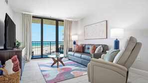 Ein Wohnzimmer mit Sofa, Sessel und Fernseher. Durch große Fenster ist der Blick auf den Strand möglich.