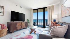 A modern living room with a flat-screen TV, a sofa, a glass coffee table, and a view of the ocean through large windows. - Ocean Trillium 604 - Beautiful Beachfront Condo in New Smyrna Beach with Oceanfront Pool (New Smyrna Beach)