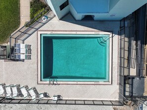 Ein Luftbild eines rechteckigen Schwimmbeckens mit einer gefliesten Liegefläche darum herum, ausgestattet mit Liegestühlen und einer Leiter.