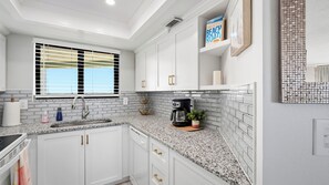 A kitchen with white cabinetry, granite countertops, a coffee maker, and a window with blinds. - Ocean Trillium 604 - Beautiful Beachfront Condo in New Smyrna Beach with Oceanfront Pool (New Smyrna Beach)