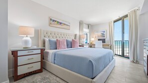 A hotel room with a large bed, a desk, a chair, a lamp, a window with curtains, and a painting on the wall. - Ocean Trillium 604 - Beautiful Beachfront Condo in New Smyrna Beach with Oceanfront Pool (New Smyrna Beach)