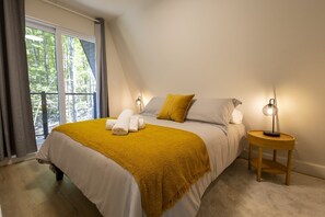 A bedroom with a bed, a bedside table, a lamp, and a view of trees through the window. - NordAstra | SPA | Accès au Lac (Chertsey)