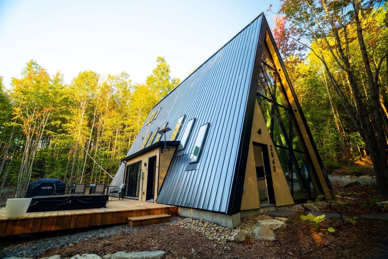 Eine moderne, dreieckige Hütte mit Metallbedachung, gelegen in einem bewaldeten Gebiet mit Terrasse und Sitzgelegenheit im Freien.