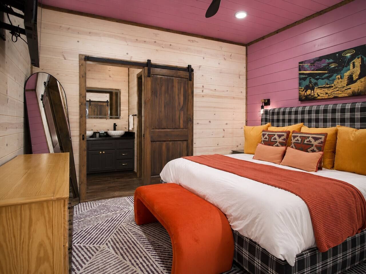 A bedroom with a bed, a bench, a dresser, a door, and a painting on the wall.