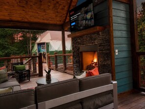 An outdoor seating area with a stone fireplace, wooden deck, and a view of a tented structure in the background. - 5BR Cabin + Glamping Dome | Sauna, ATV Trails, Pickleball (Broken Bow)