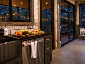 A kitchen with a large island, a sink, and a countertop with fruits and vegetables. There are cabinets with gold handles and a window with a view of trees. - 5BR Cabin + Glamping Dome | Sauna, ATV Trails, Pickleball (Broken Bow)