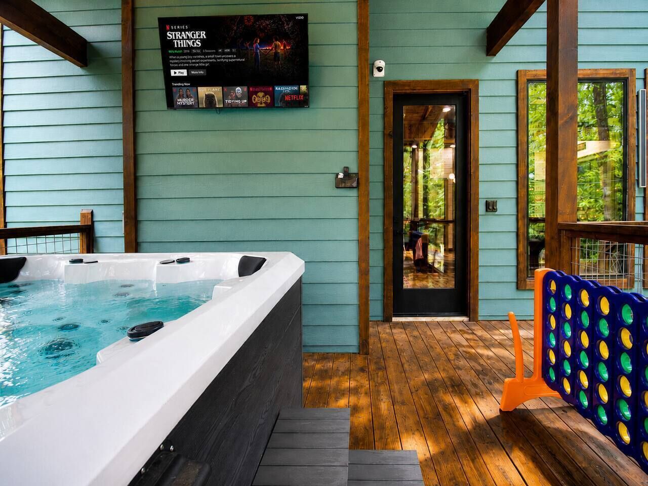 Un espace avec un jacuzzi, une terrasse en bois, un téléviseur fixé au mur diffusant une série, et une porte donnant sur un espace extérieur abrité.