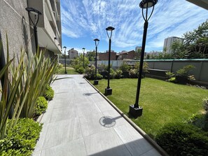 Uma passagem ladeada por postes de luz, com vegetação verde e área gramada, conduzindo a um edifício de arquitetura moderna.