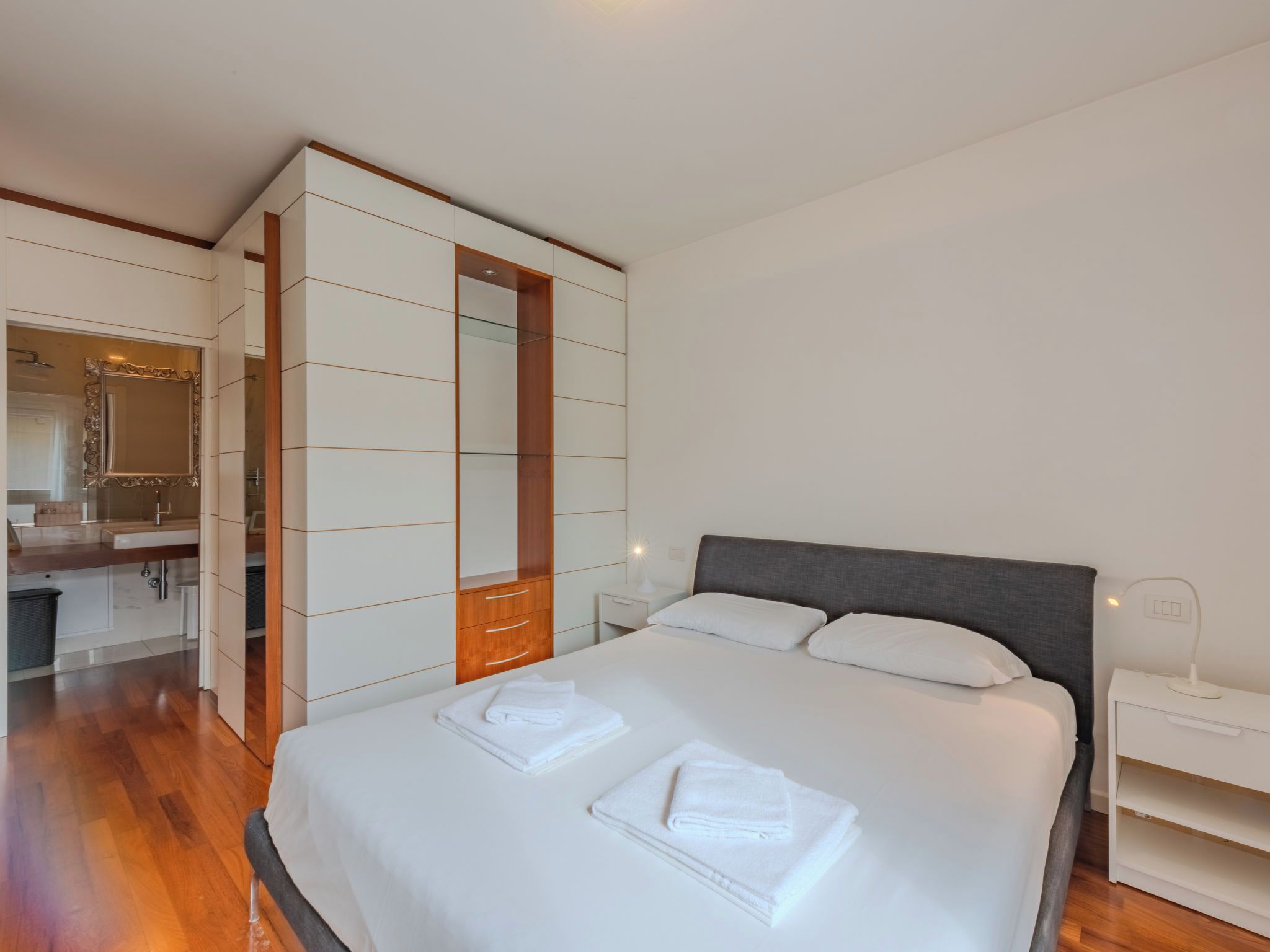 A modern hotel room with a large bed, a built-in wardrobe, a bathroom with a mirror, and a bedside table with a lamp.