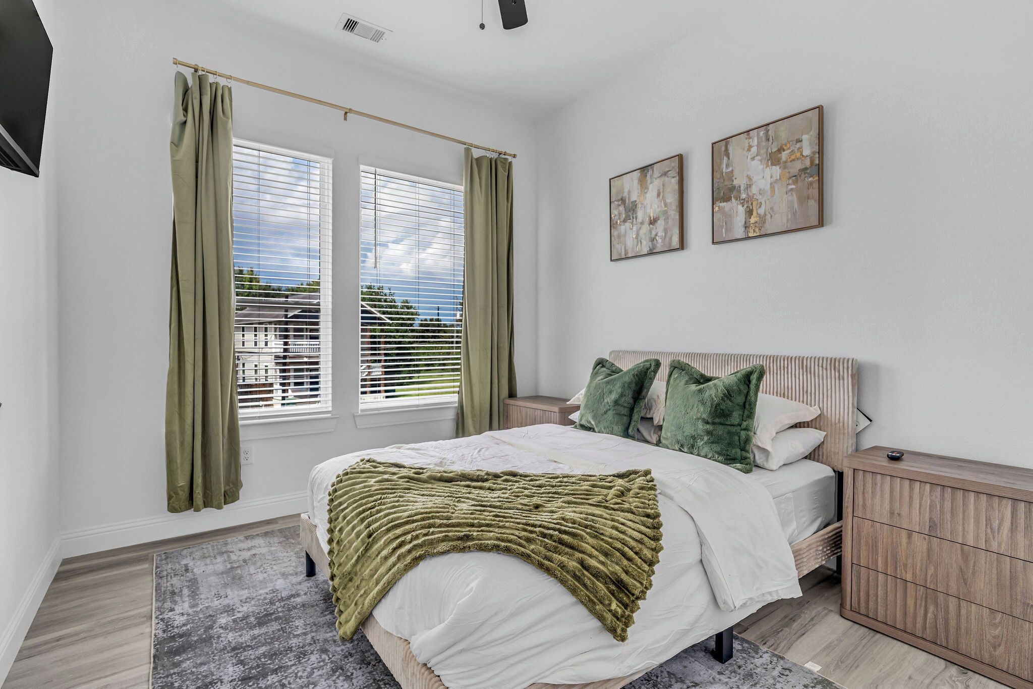 A bedroom with a bed, curtains, a bedside table, a nightstand, a headboard, and two wall-mounted artworks.