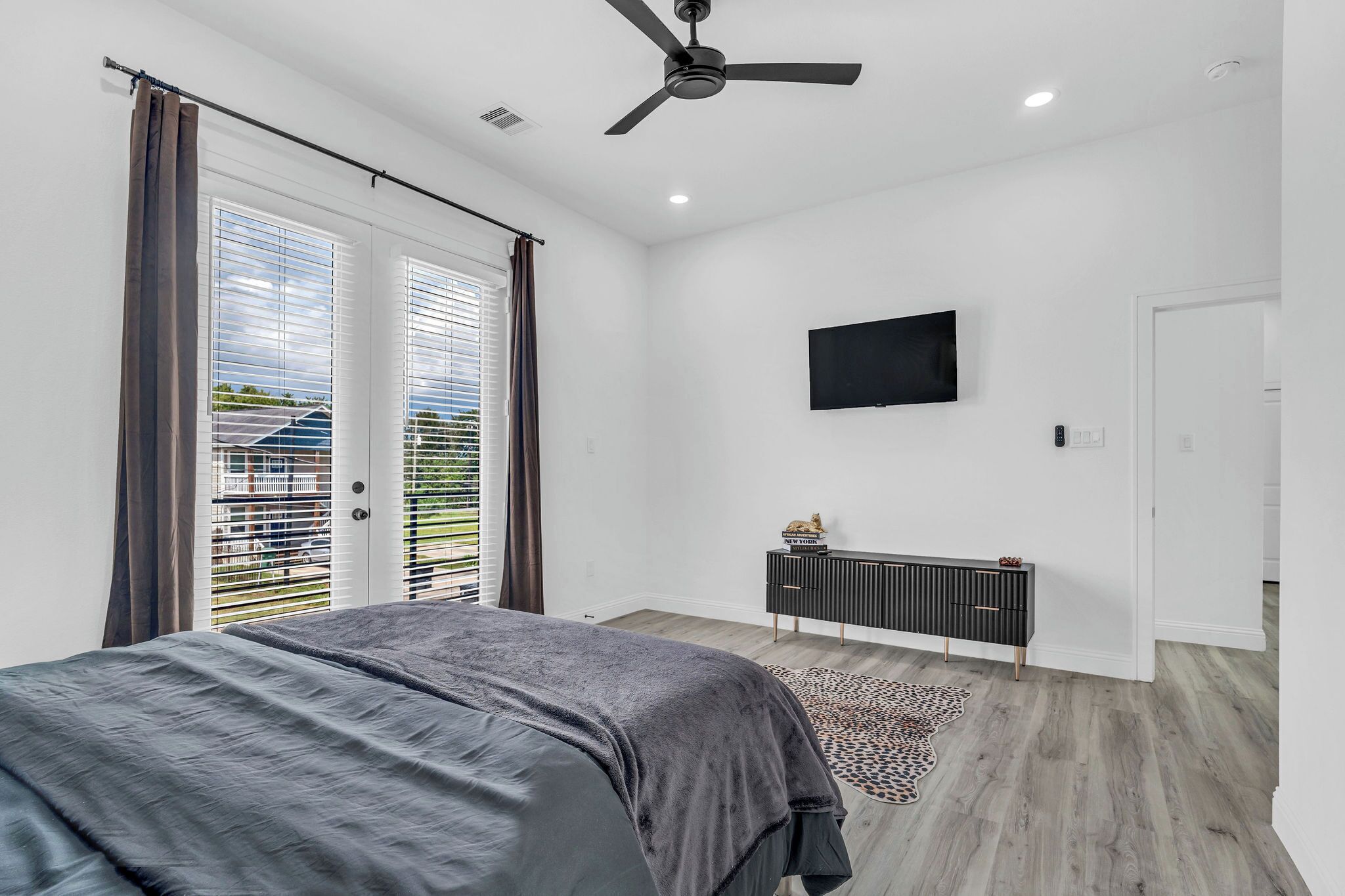 Une chambre à coucher comprenant un lit, un ventilateur de plafond, un téléviseur fixé au mur et une porte coulissante donnant sur un balcon.