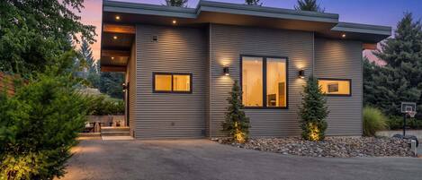 A modern two-story house with a sloped roof, large windows, and a well-lit exterior.