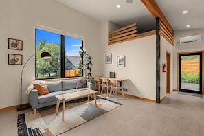 A modern living room with a sofa, coffee table, and a desk with chairs. There is a large window with a view of the outdoors.