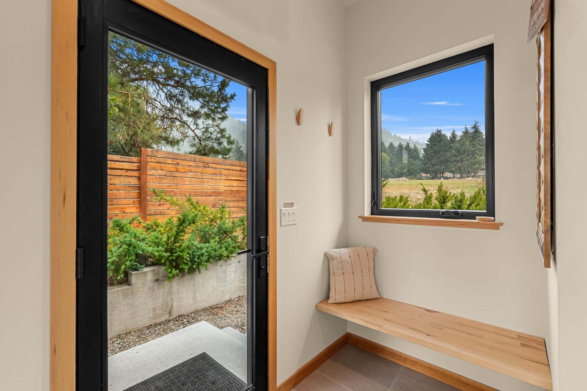 Una habitación con un banco de madera, una ventana que da a árboles y una puerta que conduce a un espacio exterior con una valla de madera.