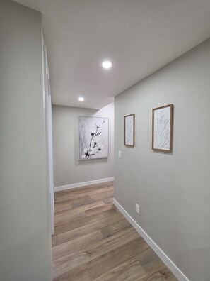 A hallway with wooden flooring, light-colored walls, and two framed artworks. - Charming 2-bedroom Basement Suite in Calgary with AC (Calgary)