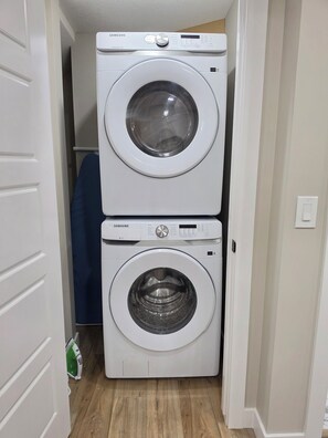 A laundry area with two stacked washing machines, a blue chair, and a green iron. - Charming 2-bedroom Basement Suite in Calgary with AC (Calgary)