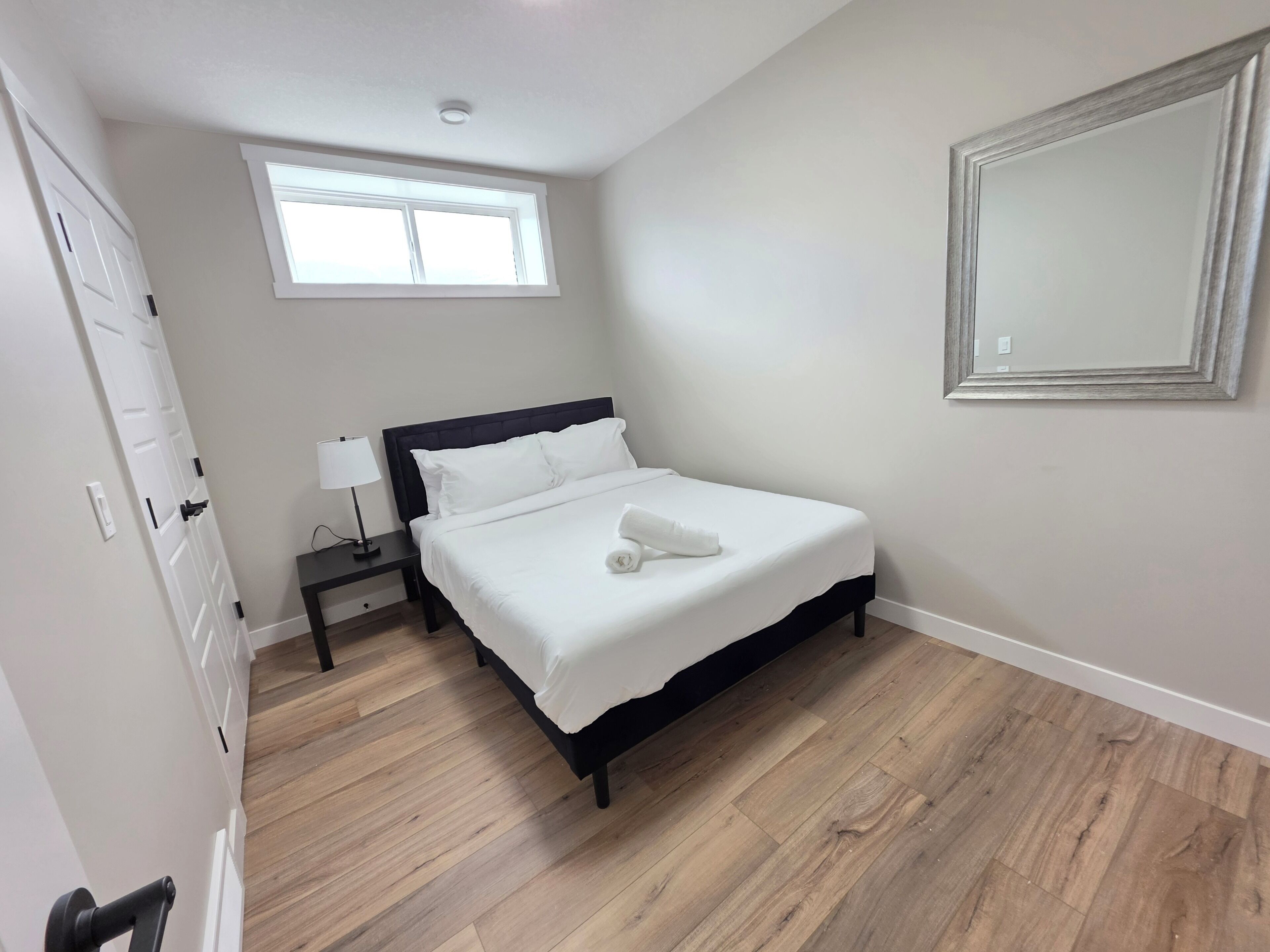 A bedroom with a bed, a nightstand, a lamp, a window, and a wall-mounted mirror.
