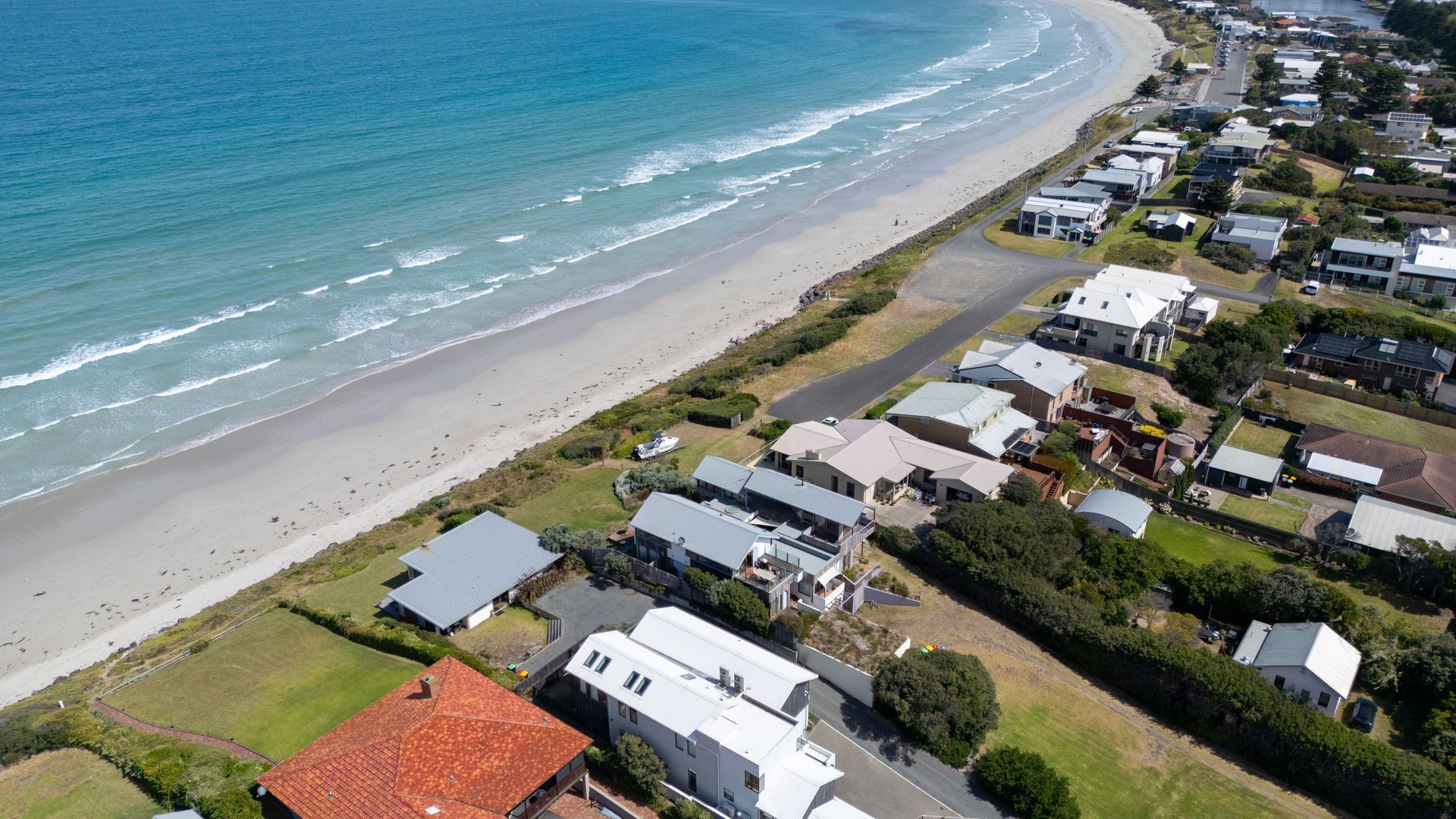 An aerial view of a coastal residential area with houses, a beach, and a road.
