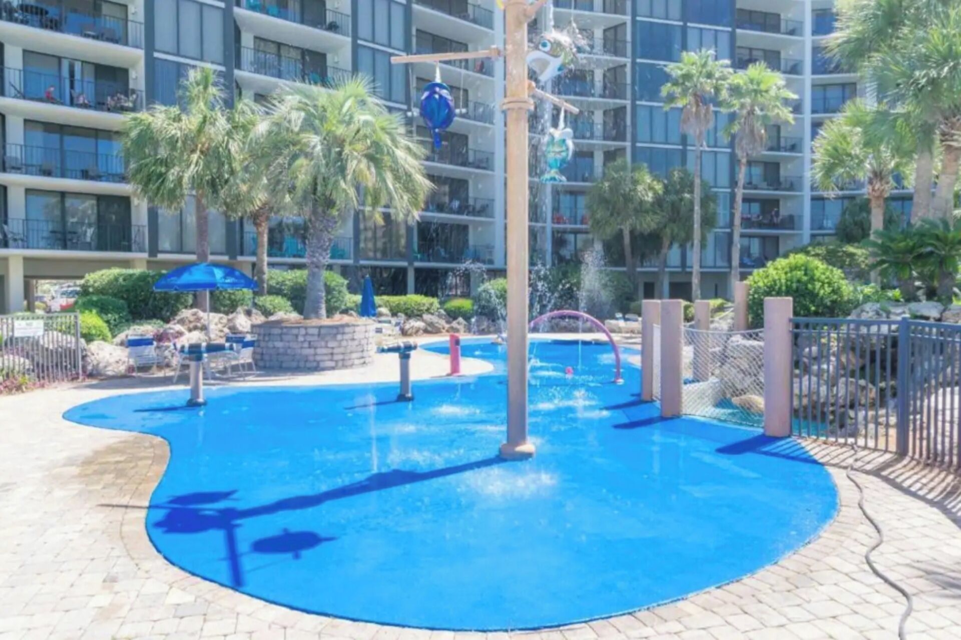 Zona de parque acuático con piscina, palmeras y un edificio al fondo.
