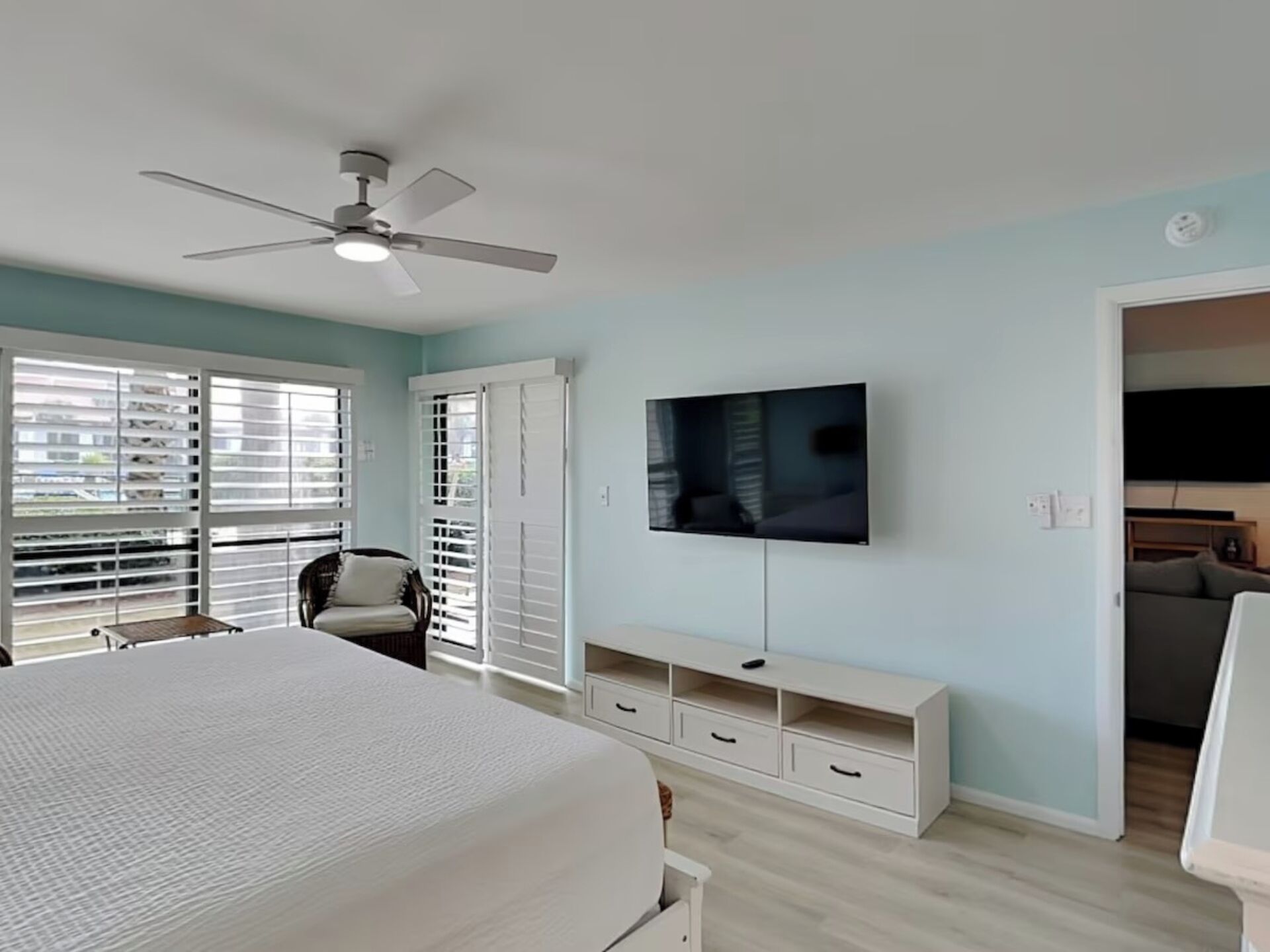 A bedroom with a bed, a TV mounted on the wall, a ceiling fan, and a window with blinds.