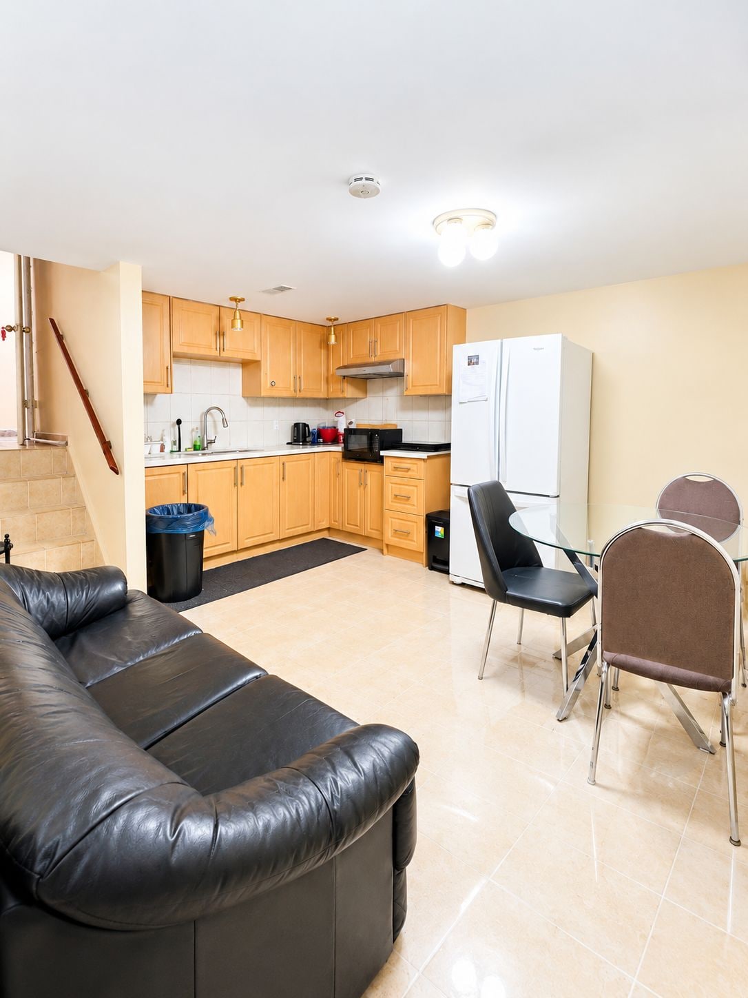 Una sala de estar moderna con un sofá de cuero, una mesa de comedor con sillas, una zona de cocina con armarios de madera, un refrigerador blanco y un cubo de basura.