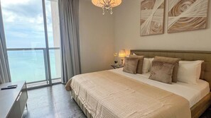 Quarto de hotel com uma cama grande, uma mesinha de cabeceira, um abajur e vista para o oceano pela janela.