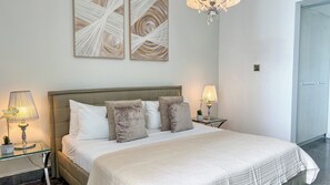 Uma cama bem arrumada com edredom branco e duas almofadas, com mesas de cabeceira com luminárias e um vaso de planta.