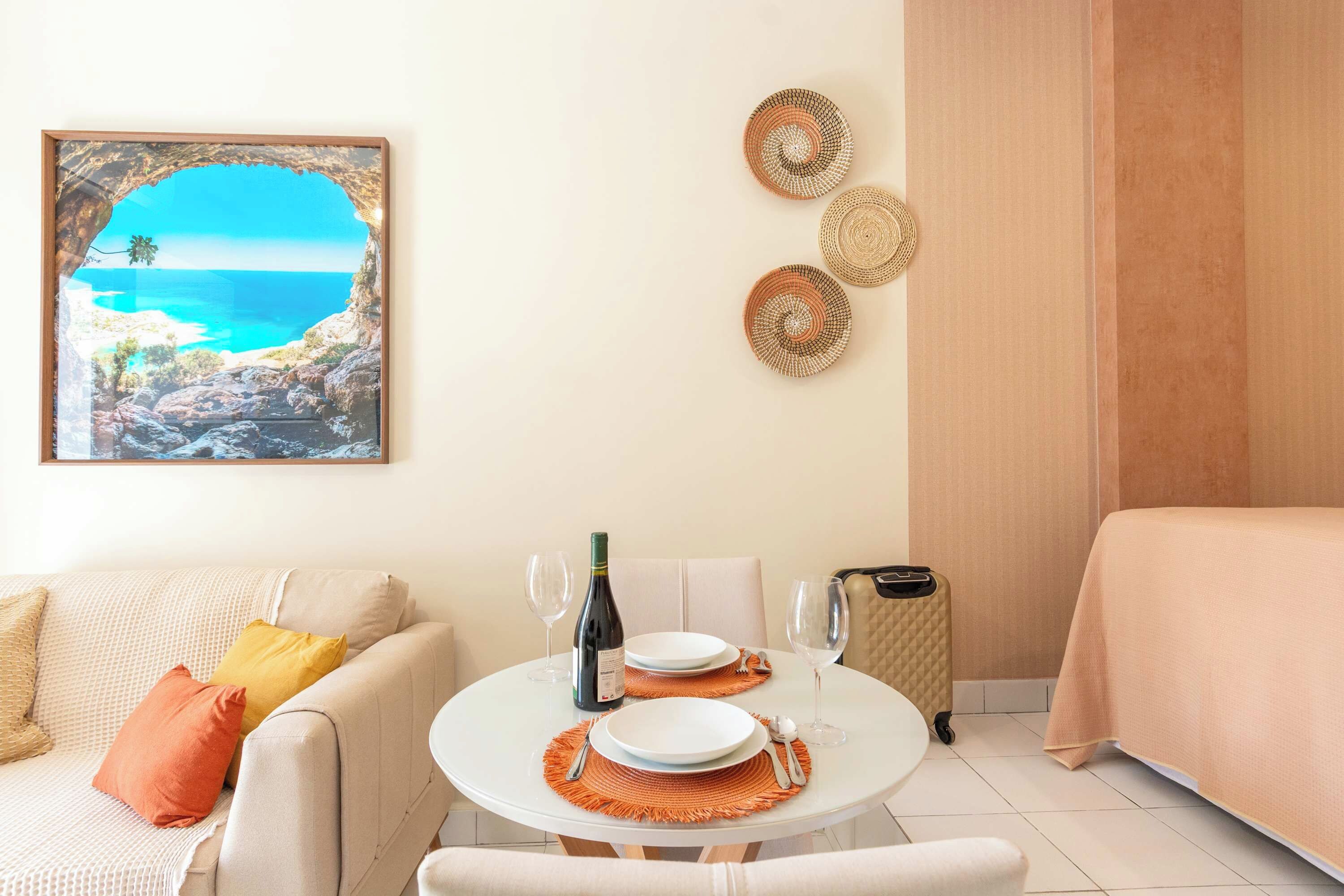 Quarto com uma mesa de jantar redonda posta para dois, um sofá, uma cama e um quadro com paisagem costeira emoldurada.