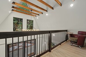 Une chambre spacieuse aménagée en mezzanine, avec un plafond haut, des poutres apparentes et une vue sur l’extérieur grâce à de grandes fenêtres.