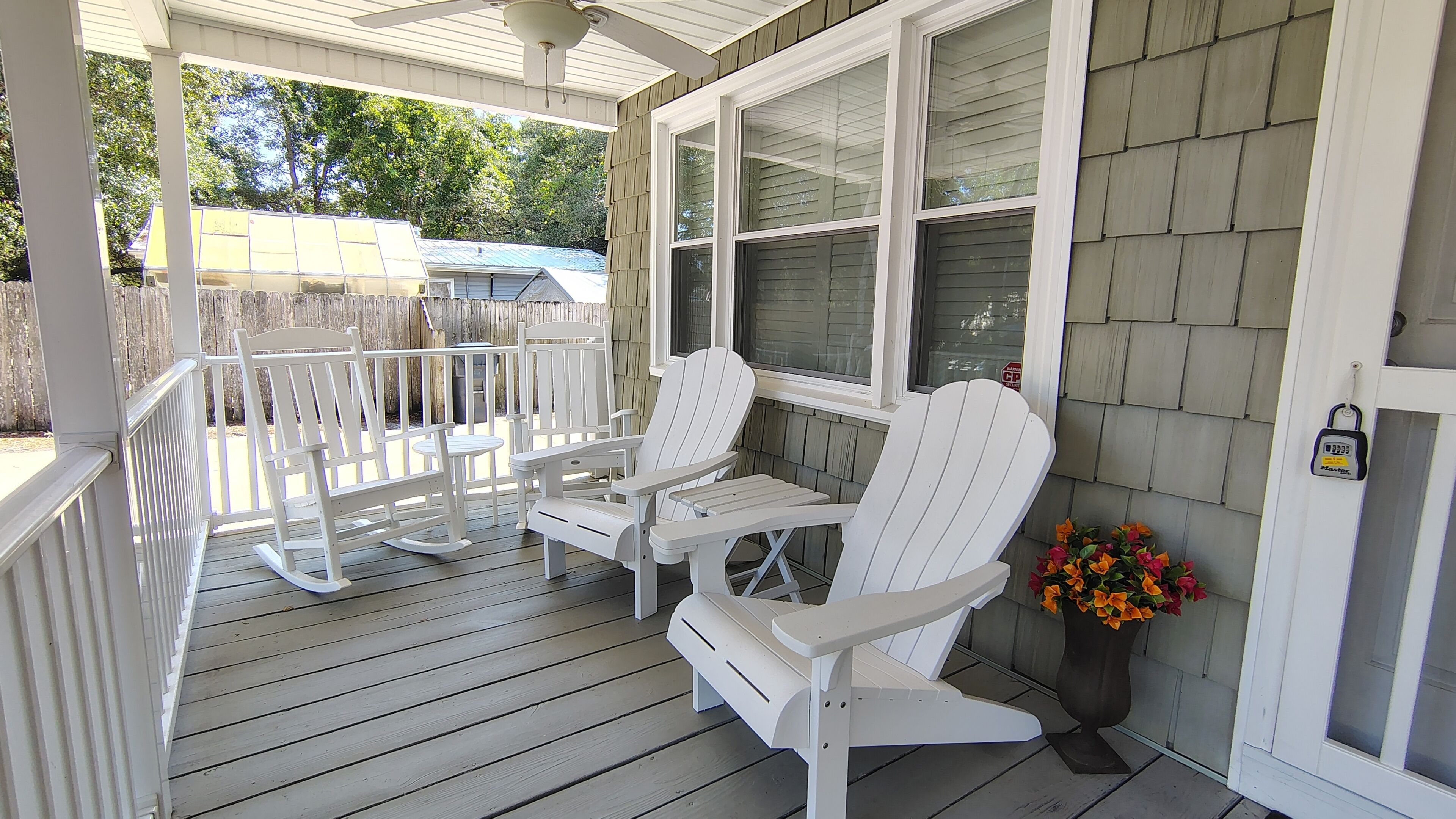 A covered porch with white Adirondack chairs and a table, a wooden fence in the background, and a potted plant with flowers.