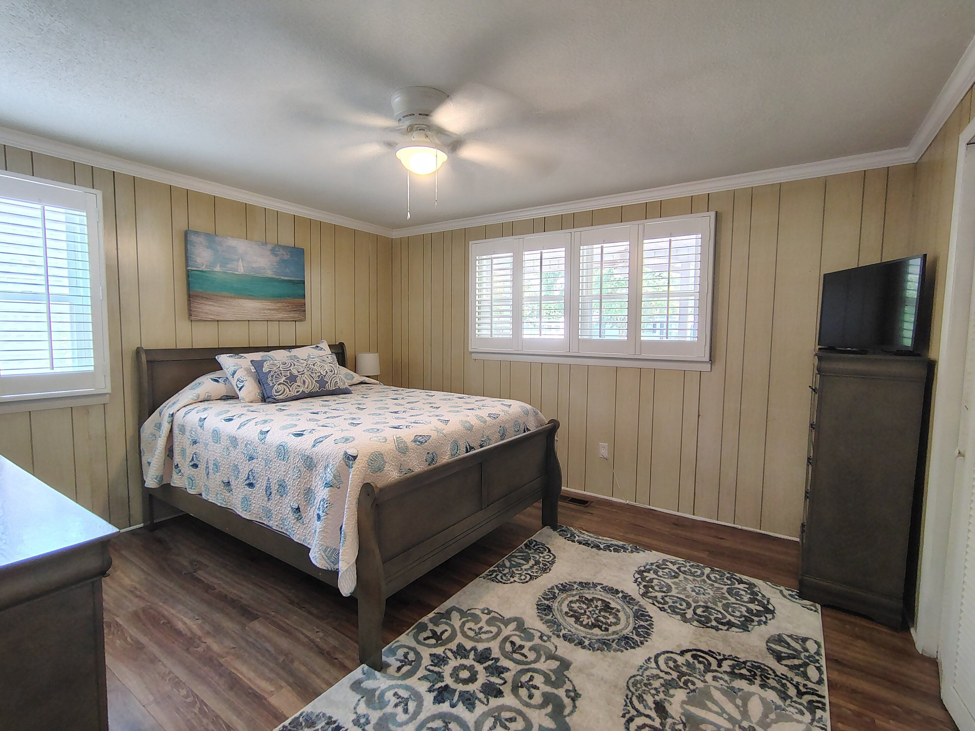 A bedroom with a bed, a painting, a ceiling fan, a window with blinds, and a television on a stand.