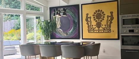 A modern dining area with a glass table and metal chairs, a large abstract painting, and a kitchen with Bosch appliances.