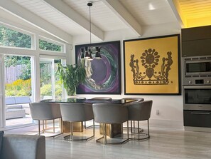 Ein moderner Essbereich mit einem Glastisch und Metallstühlen, einem großen abstrakten Gemälde und einer Küche mit Bosch-Geräten.