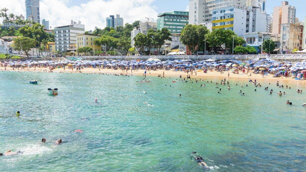 Uma praia com águas cristalinas, pessoas nadando e tomando sol, e um horizonte urbano ao fundo.