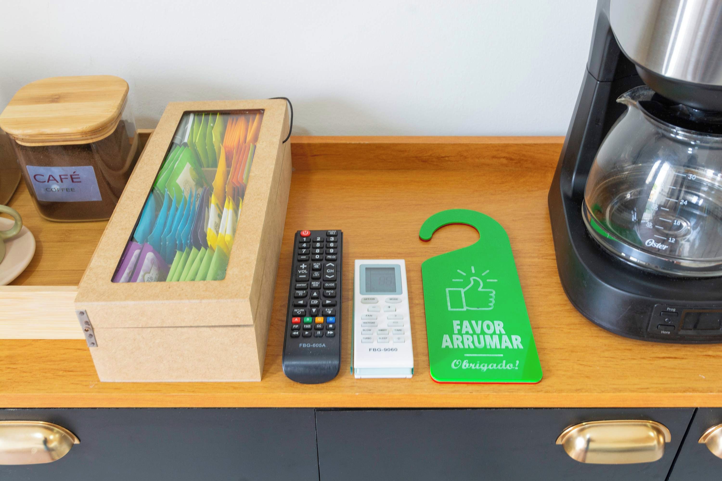 A kitchen counter with a coffee maker, a jar of coffee, a box of colorful pencils, a remote control, a digital clock, and a green tag with 'Favor Arrumar' written on it.