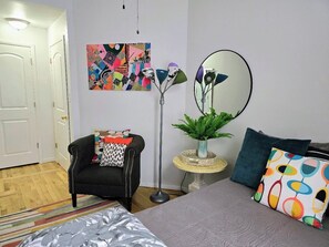 Une pièce comprenant un lit, une chaise, un miroir rond, un tableau coloré et une petite table sur laquelle est posée une plante.