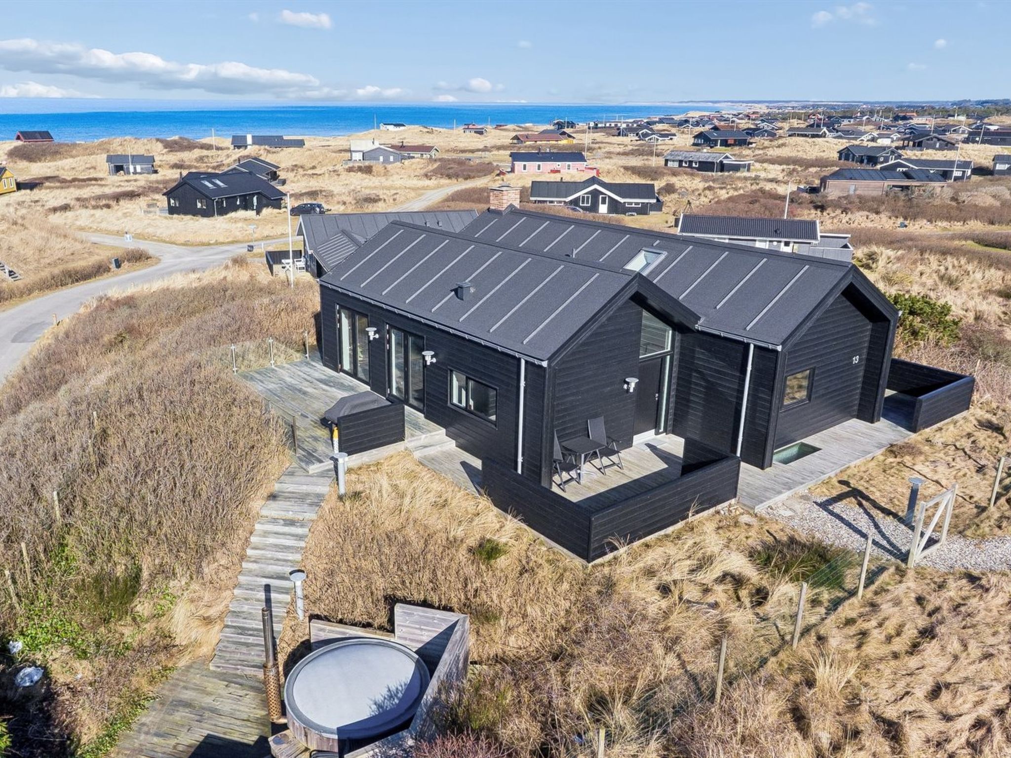 Un bâtiment moderne de couleur noire, doté d’un toit plat, situé dans une zone côtière, avec des dunes et une plage en arrière-plan.