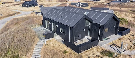 Un edificio moderno de color negro con techo plano, situado en una zona costera con dunas y playa al fondo.