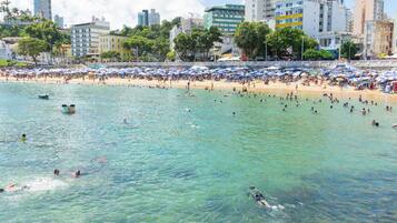 A beach with clear water, people swimming and sunbathing, and a cityscape in the background.