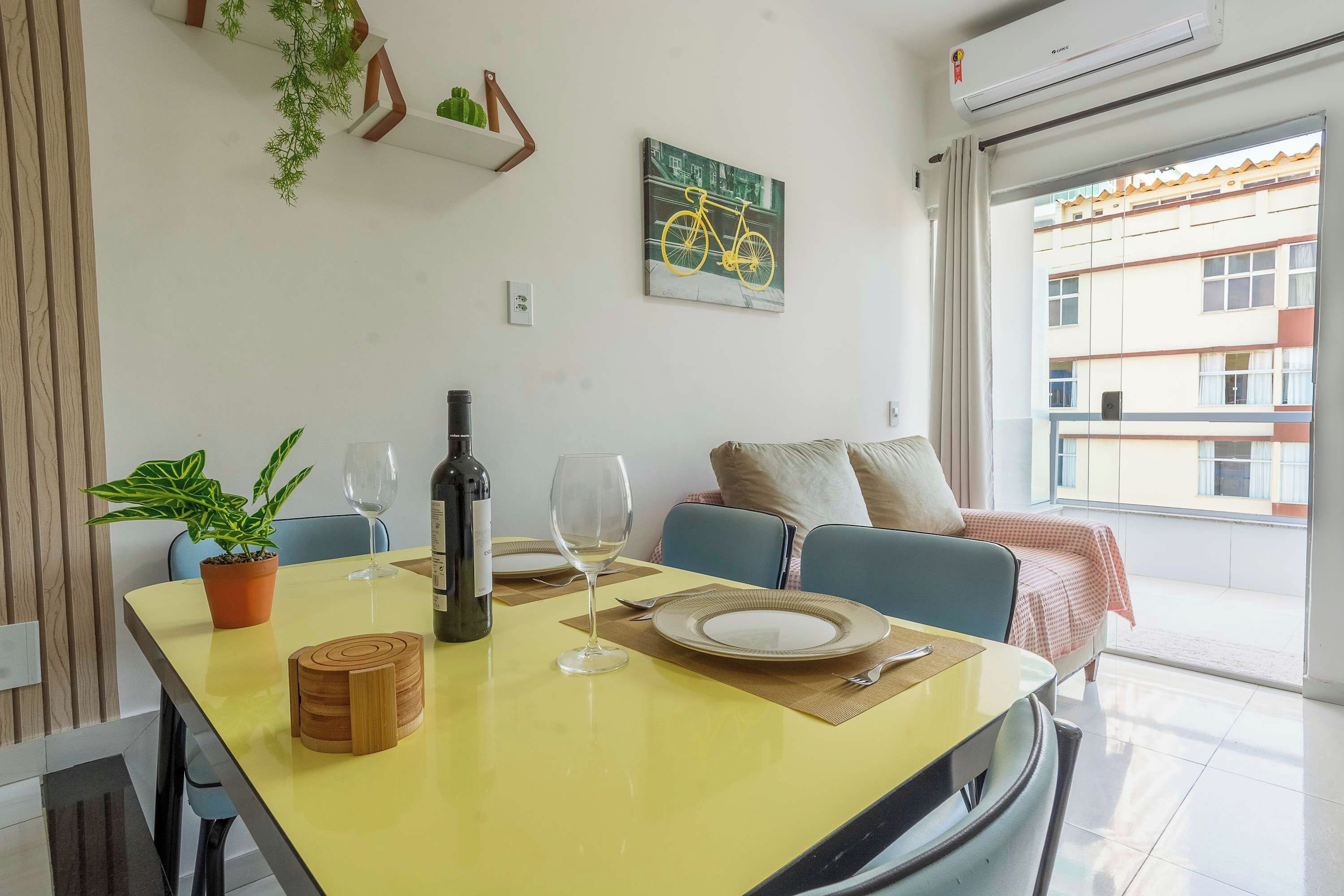 Un coin repas avec une table jaune, un verre à vin, une bouteille et une assiette. On y trouve également un canapé bleu et une fenêtre donnant sur des immeubles.