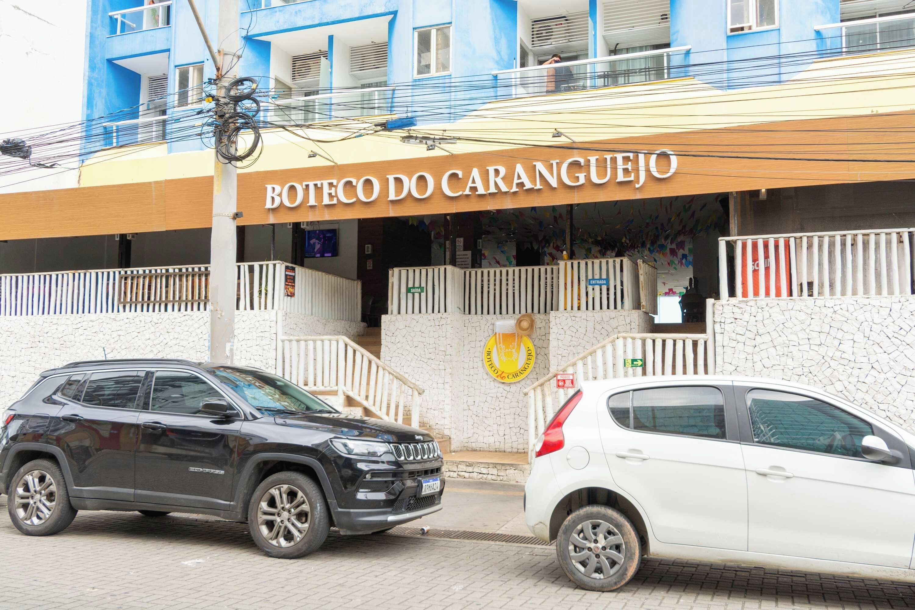 Un edificio con un letrero que dice 'Boteco do Caranguejo' y un logotipo con una caracola. Hay dos coches estacionados adelante.