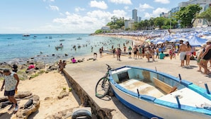 Uma praia com uma fileira de barcos, uma bicicleta e pessoas aproveitando a água.