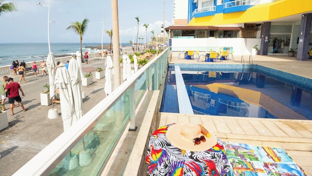 Área de piscina com vista para o oceano, um edifício com varandas e um calçadão à beira-mar.