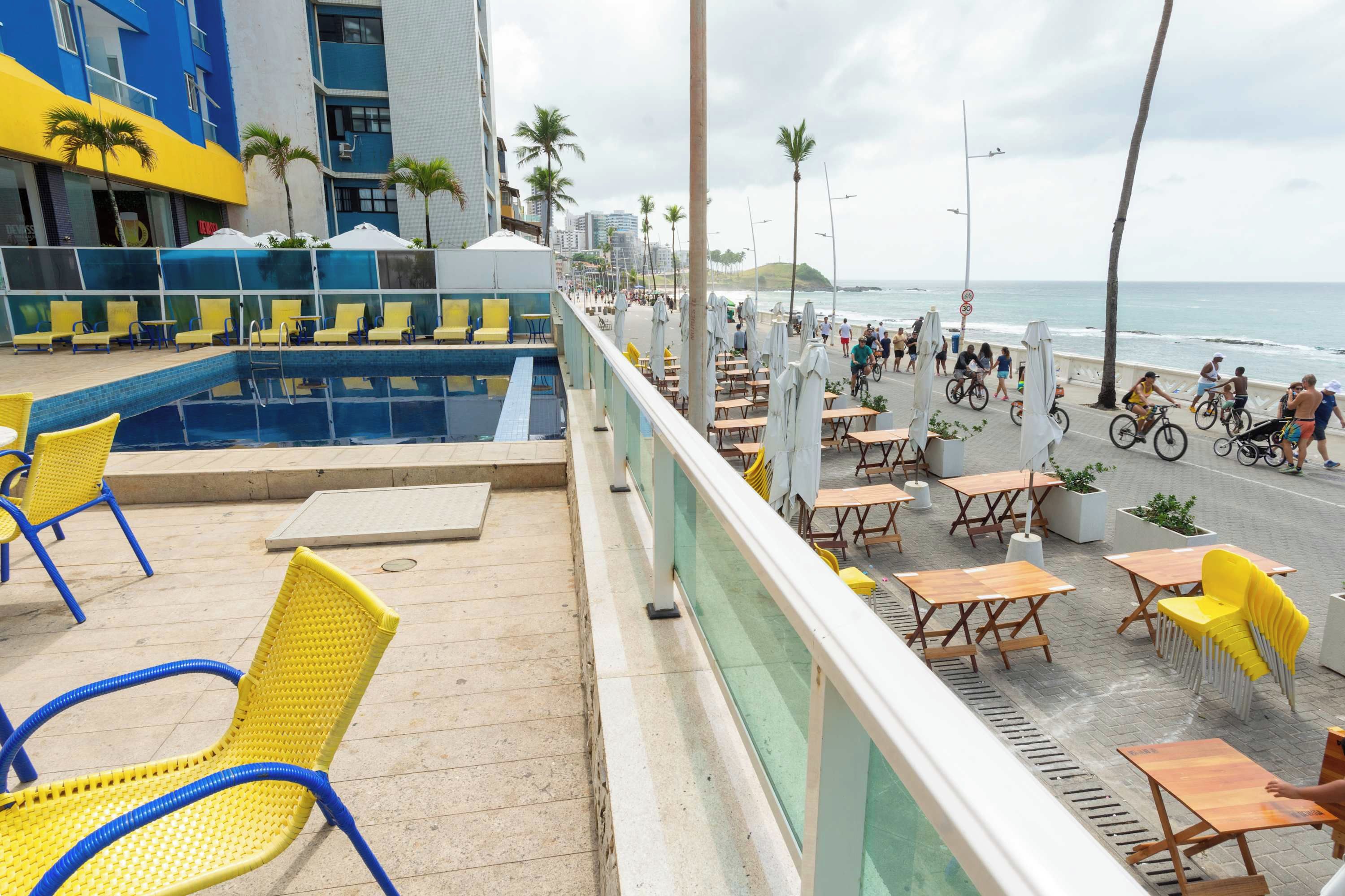 Área da piscina com cadeiras amarelas e azuis, piscina e vista para a praia e o oceano.