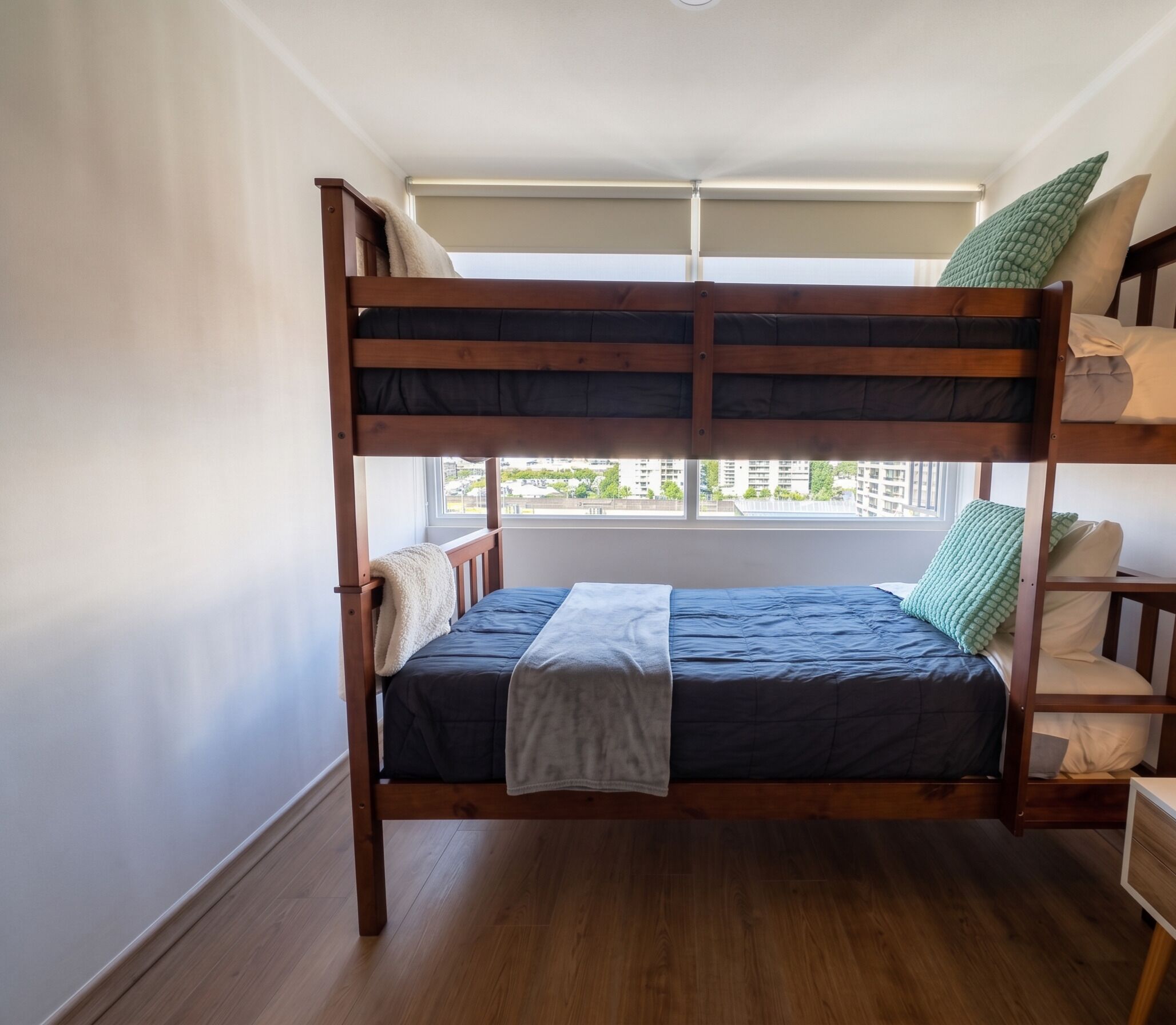 Cama individual superpuesta con ventana, suelo de madera y vistas a edificios.