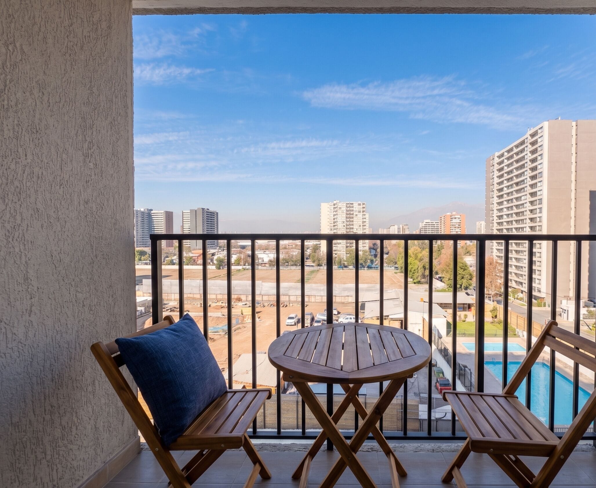 Un balcón con una mesa de madera y dos sillas, con vistas a una piscina y al paisaje urbano.